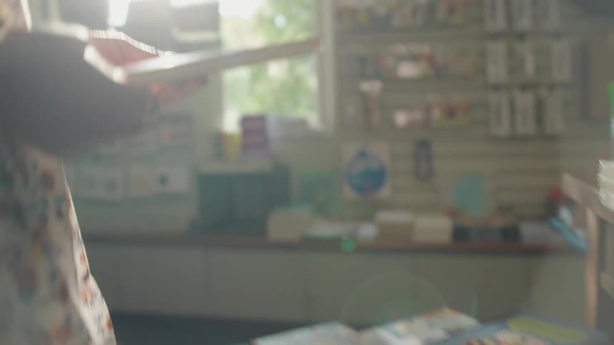 A person with a floral dress and a yellow scrunchie on their wrist is stacking small books or notepads on a wooden table likely in a stationery or book storethe camera focuses on their hands as they handle a stack of books with red spines and a plain