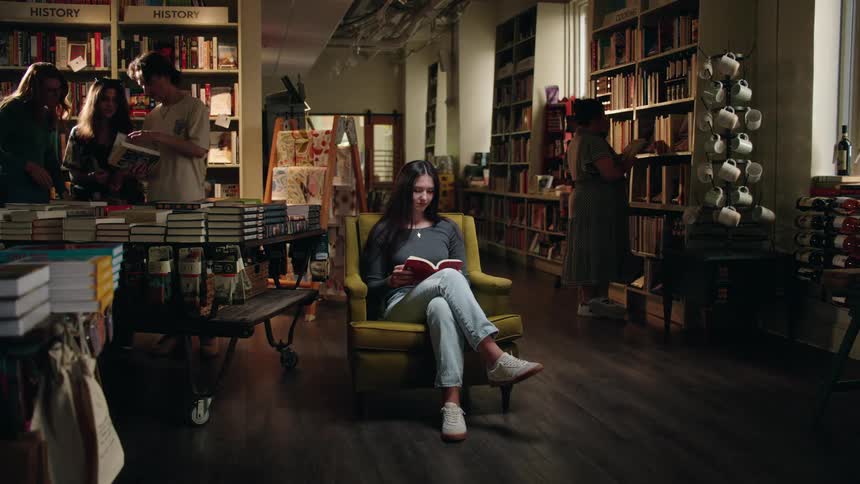 Teenager girl sitting on an armchair at a library reading a book surrounded by other people interacting with books indoors during day time