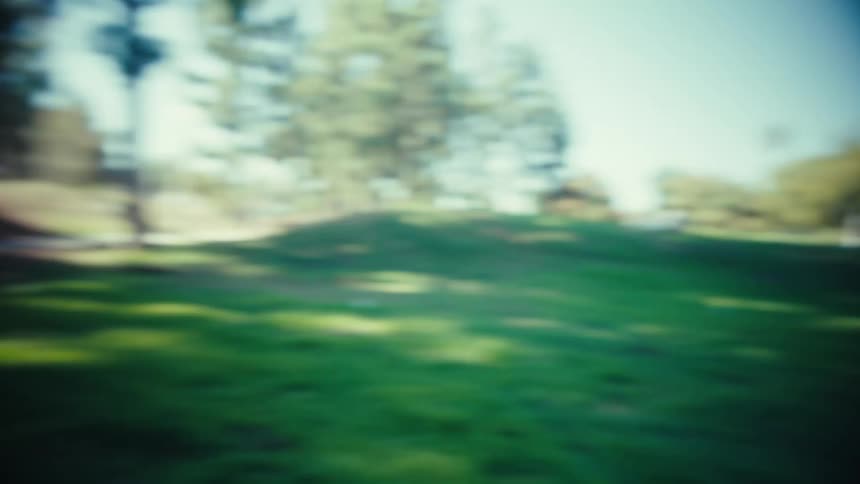 A smiling man in a baseball jersey grabs the camera to pan across a grassy park field before turning the lens back onto himself for a closeup