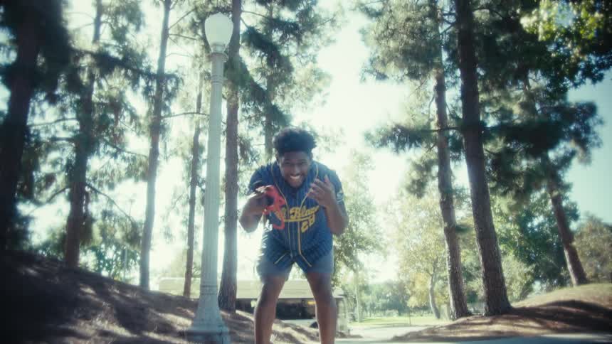 A young man wearing a baseball jersey and shorts stands on a paved pathway in a sunny park surrounded by tall trees and a lamp post he is smiling and enthusiastically holding a pair of red boxing gloves in his hands flexing his arms