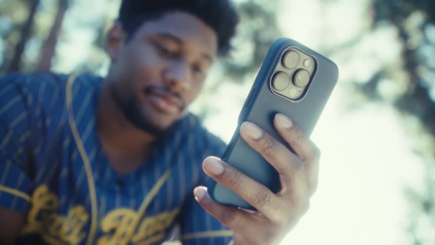 Man wearing a blue sports jersey holding a blue smartphone in an outdoor area