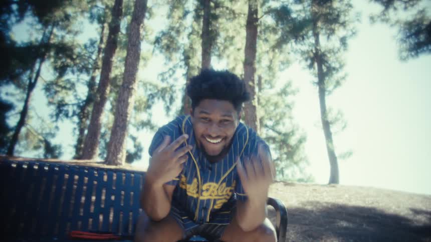 A young man wearing a blue and yellow pinstriped baseball jersey sits on a park bench he starts by looking pensive then smiles and leans into the camera