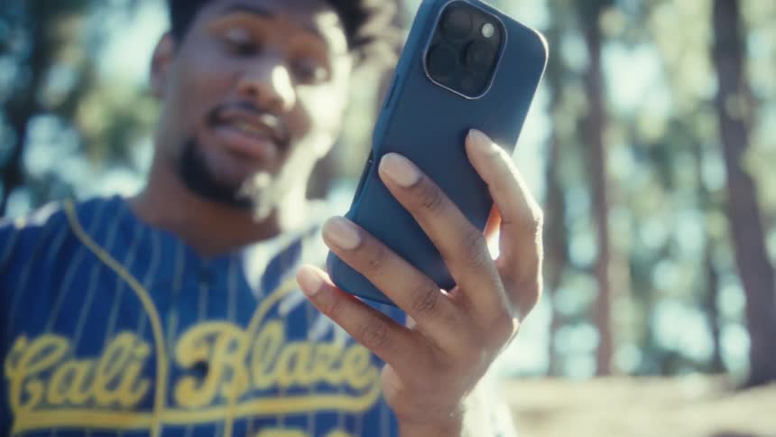 A young man wearing a striped baseball jersey holds a dark smartphone in a blue case with his right hand with the entire scene brightly lit and set against a heavily blurred backdrop of green trees and bright daylight