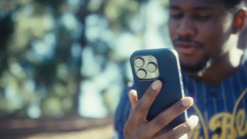 A black man in a baseball jersey is looking forward talking focused on the screen of the mobile phone he is holding outdoors