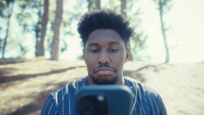 A man with dark textured hair and a beard wearing a pinstriped shirt sits outdoors and intently looks down at the screen of a smartphone he holds to take a selfie