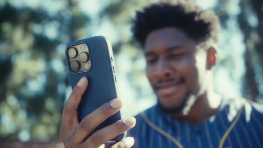 A closeup focuses on a mans hand holding a smartphone in a dark case with the mans smiling face visible in the background while he views the screen outdoors beneath blurred trees
