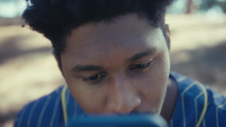 Young black man sitting on a bench at a park looking very closely at the screen of a smartphone outdoors during day time