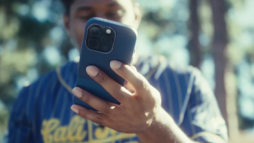 The video is a closeup shot focused on a persons hands holding a darkcased smartphone outdoors the person who appears to be a young man wearing a blue and yellow striped baseballstyle jersey is scrolling or interacting with the phone smiling and l