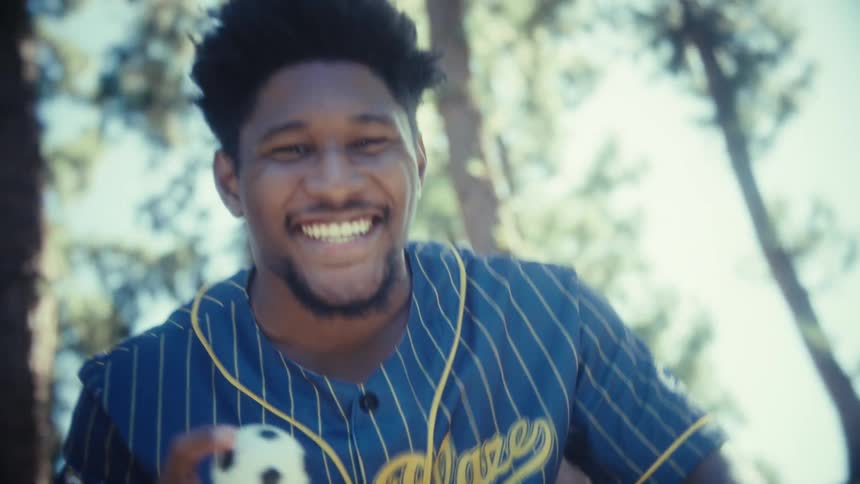 Man in blue and yellow striped shirt holds spotted ball in wooded area under sunlight