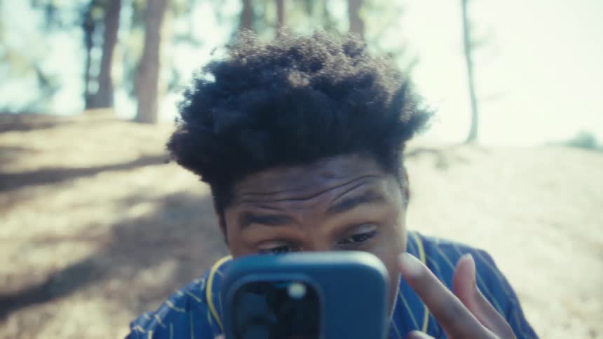 A young man holds up a smartphone to check his reflection and touches his face while outdoors