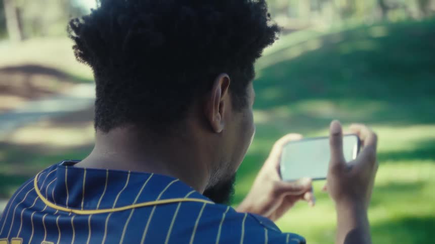 A young black male wearing a sports jersey with the number 28 and the name player on the back sits on a bench in a public park at midday holding a mobile device and watching a game with excitement