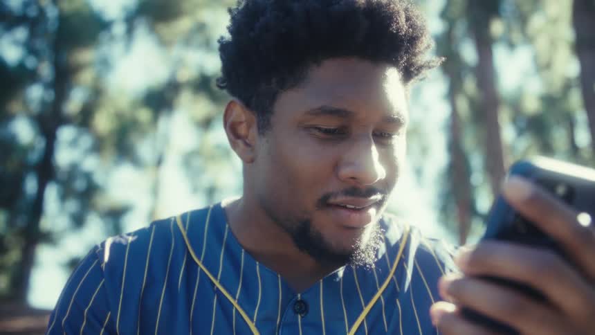 A black man is outdoors talking to his smartphone that he is holding while smiling