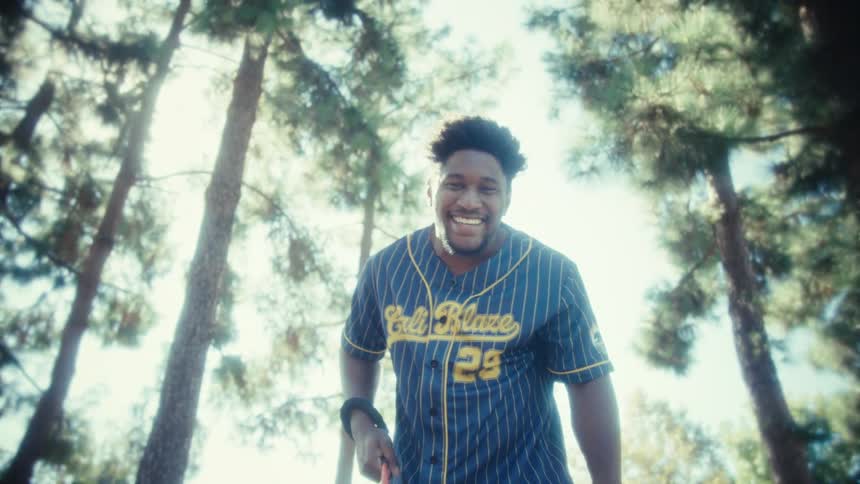 A young black man wearing a jersey walks his dog with excitement through a public park surrounded by trees at midday