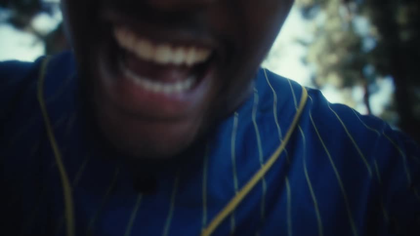 A young man wearing a blue and yellow cali blaze 28 baseball jersey runs toward the camera on a paved path lined with tall trees he is smiling excitedly and the shot finishes in a closeup of his broadly grinning face with the bright sky and trees blu