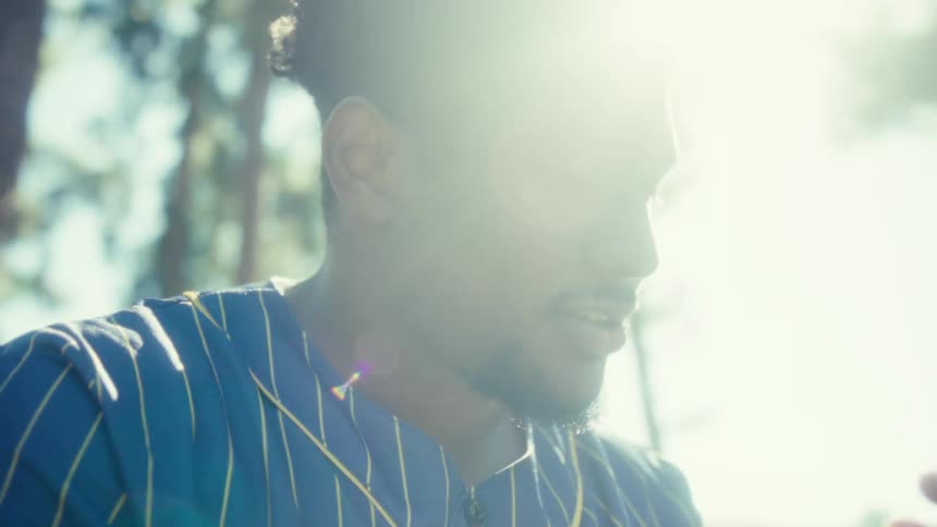 A young man sits on a public bench in a park making a video call on his phone