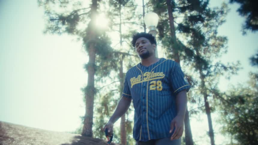 A man wearing a blue and yellow baseball jersey walks down a sunny treelined slope in a park holding a red leash in his hand
