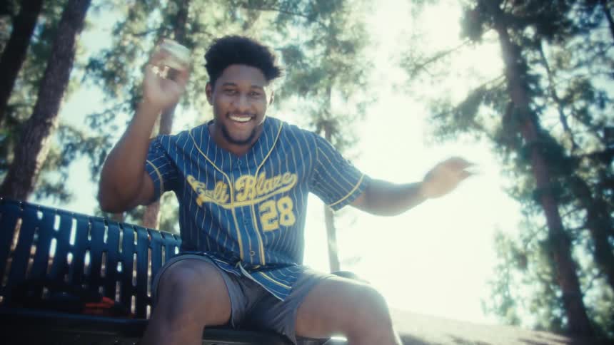 A young man wearing a striped baseball jersey is sitting on a park bench looking at his phone and smiling before he playfully holds up and then throws a small soccer ballthe camera follows the balls trajectory as it rolls down a bright grassy hill