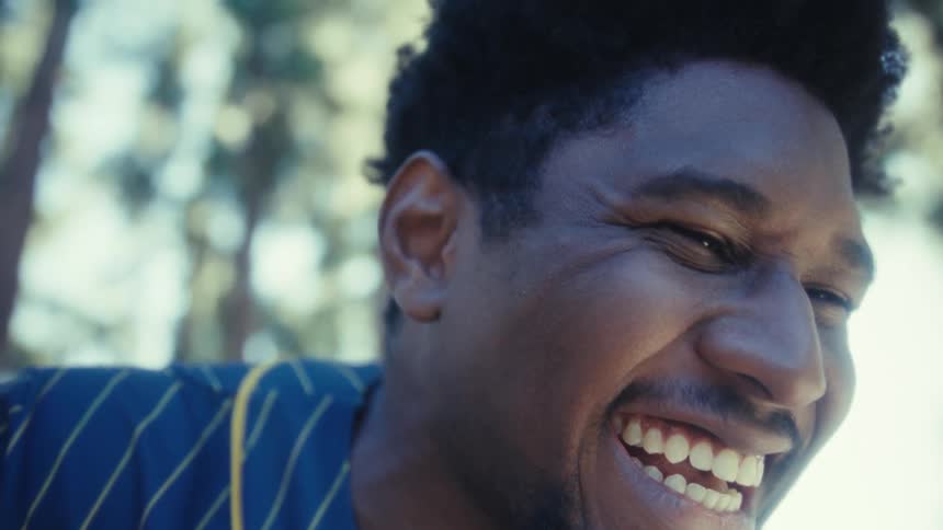 A man wearing a blue and yellow pinstripe baseball jersey is seen outdoors under bright sunlight looking down at his smartphone before bursting into a wide laugh and smile in reaction to what he is viewing on the screen