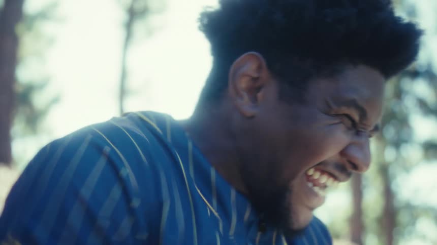 Sitting in a sunlit park a young man wearing a baseball jersey reacts with shock and then hearty laughter while looking at his smartphone