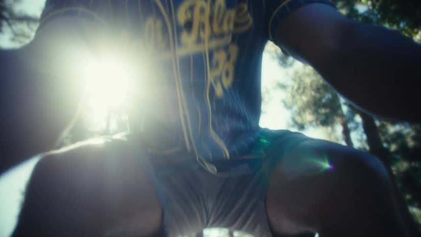 A man wearing a blue and yellow pinstripe baseball jersey is first seen running playfully down a path in a heavily wooded park and then moves in for an extremely close wideangle shot smiling broadly into the camera lens