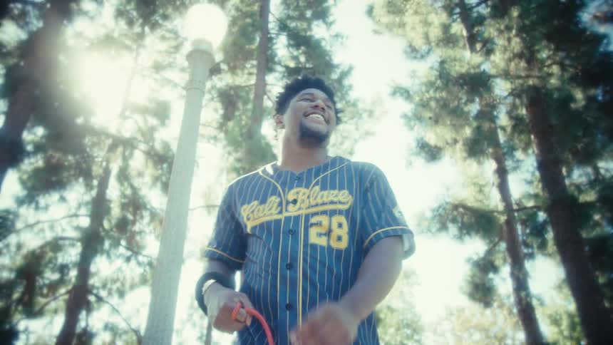 A man wearing a blue pinstripe baseball jersey stands in a sunny park with tall trees overhead initially looking around before smiling and gesturing directly toward the camera while holding a red leash