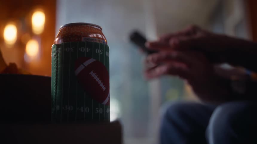 A close view of a soda can wearing a football themed koozie sits on a table in a dark room with a bright light flaring behind it and a person using a remote control in the blurred background