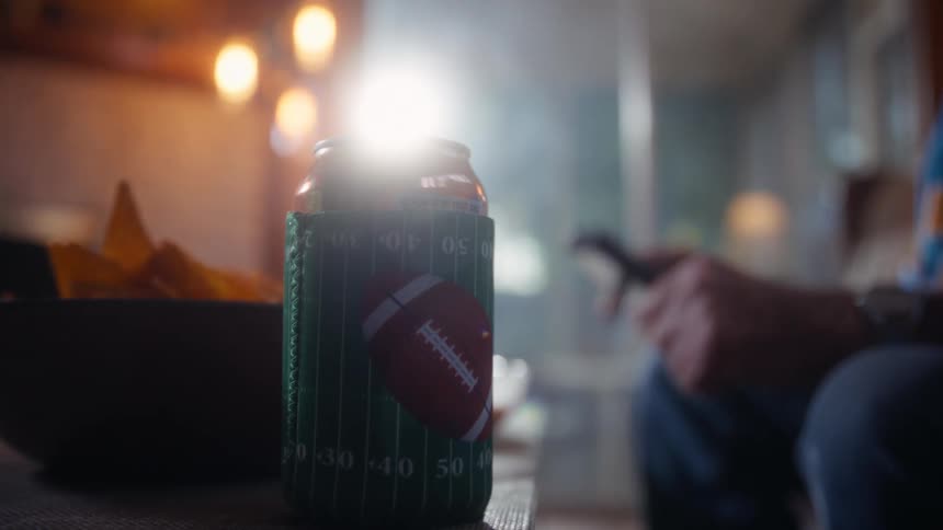 Can wrapped in an american footballthemed cover beside a bowl of snacks as someone holds a remote control
