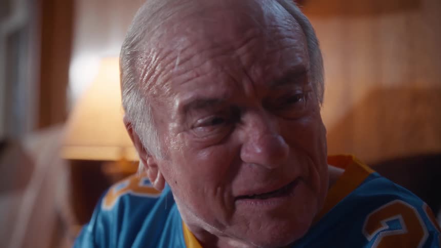A concerned elderly man in a football jersey turns to console his distressed wife as they watch an intense game together with deep emotion