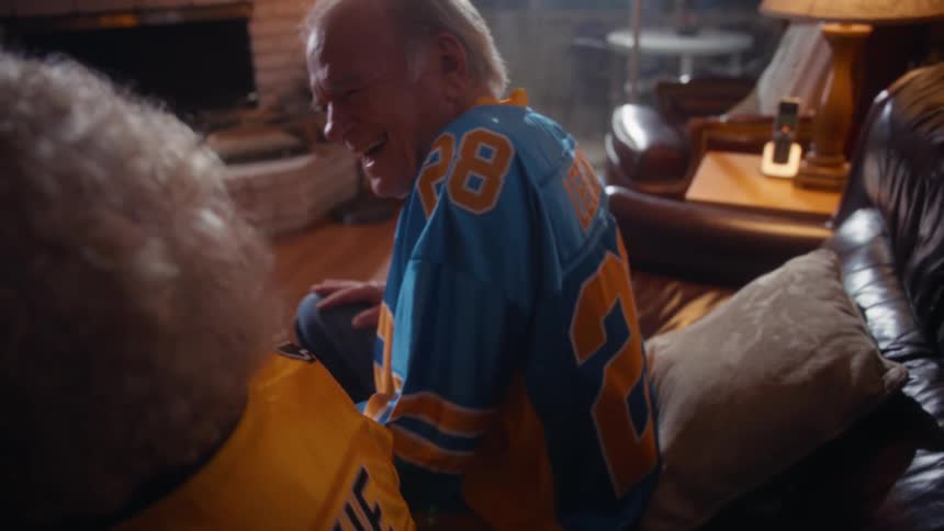 A short closeup view shows an elderly couple sitting on a couch wearing blue and yellow sports jerseys with the womans jersey having rookie above the number 7 the man is laughing heartily suggesting a happy or amusing moment while watching a game