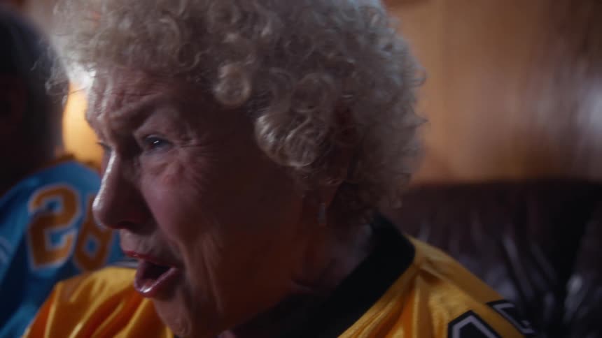 Elderly woman and elderly man wearing football jerseys cheering and clapping in a living room indoors during night time