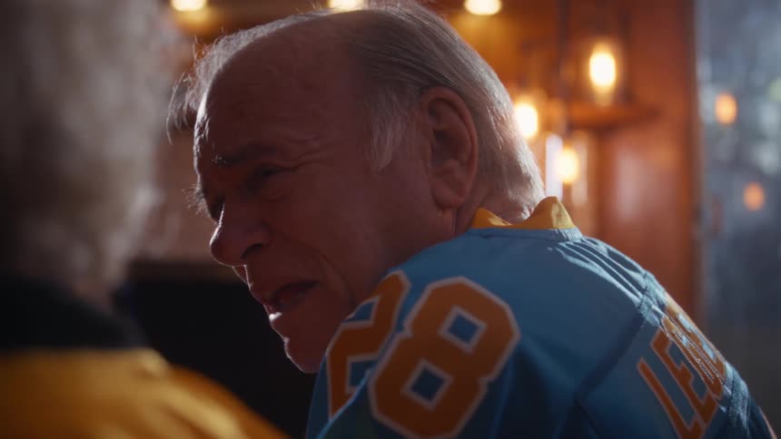 Elderly man and woman wearing football jerseys and sitting in the living room after dusk speak to each other