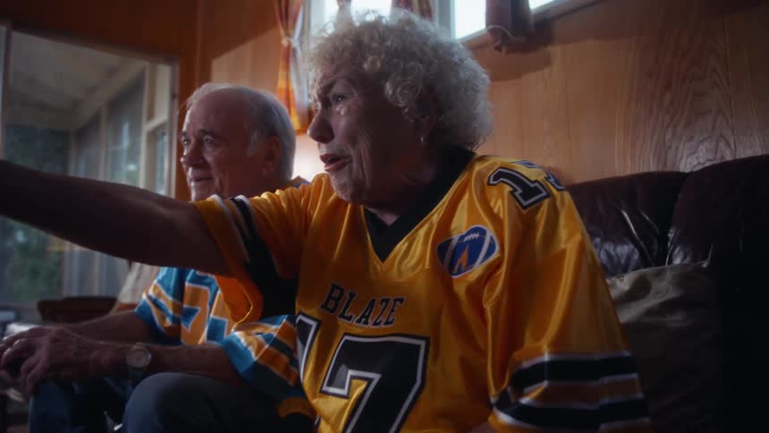 In this video an elderly couple is enthusiastically watching a game on television in a woodpaneled living roomthey are seated on a leather couch both wearing colorful football jerseys the woman in a yellow blaze jersey with the number 17 the ma