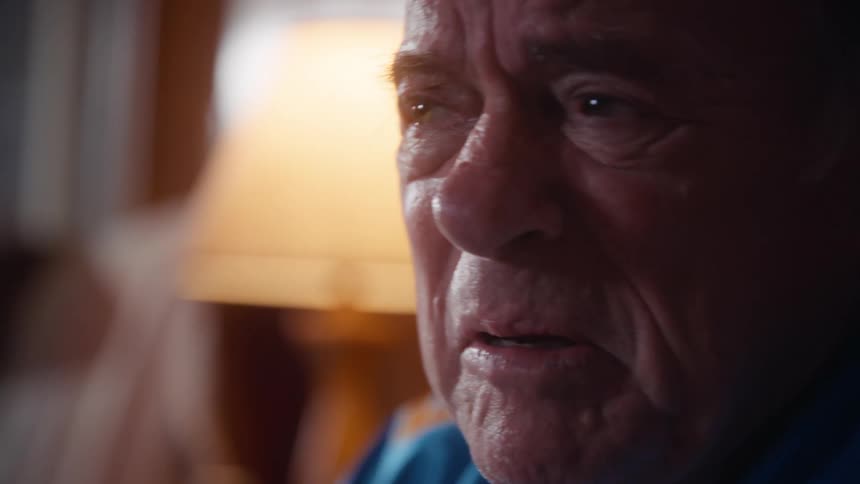 The profile of an elderly man wearing a blue and yellow sports jersey who appears deeply tense as he stares intently offcamera with soft light illuminating his face from the side