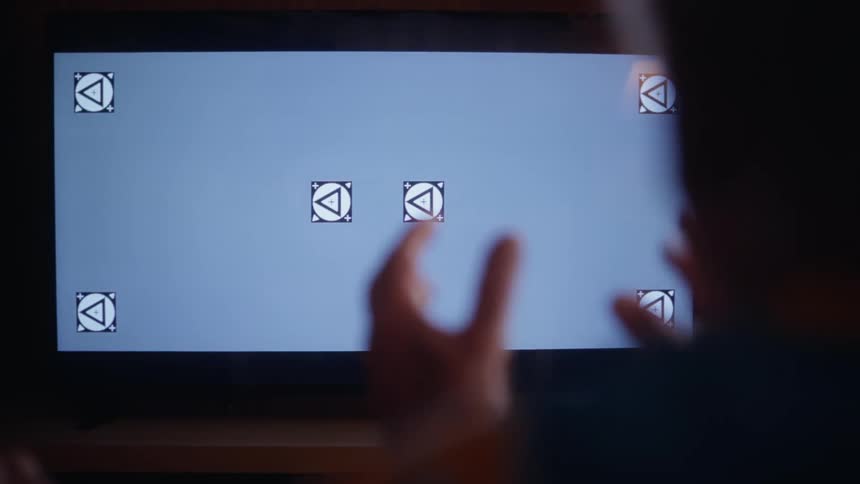 The video shows a persons hands touching a television screen displaying a gray background with several triangular tracking markers in a calibration or testing setup the person in the foreground is blurred indicating a close focus on the screen and the