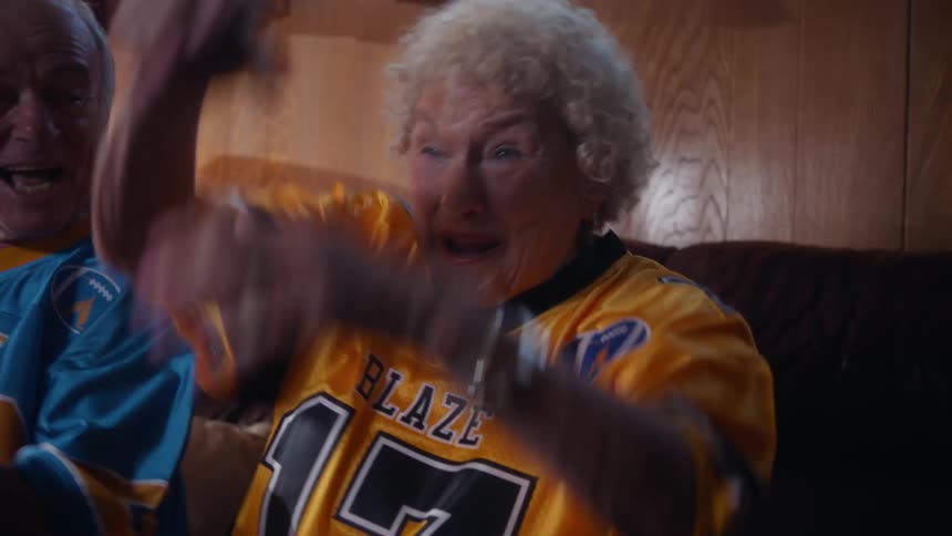 An excited elderly couple wearing matching blaze football jerseys in yellow and blue watch a game on tv from their couch they cheer enthusiastically with the woman celebrating a big play by throwing her hands up and smiling broadly
