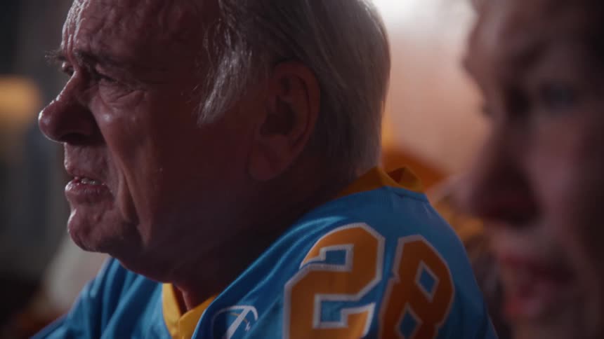 An elderly couple sits on their living room couch wearing rugby sports jerseys filled with sadness and disappointment