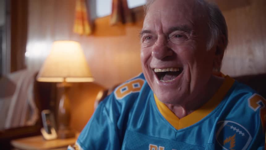 Elderly man and elderly woman talking while cheering and laughing wearing football jerseys sitting on a couch indoors during day time