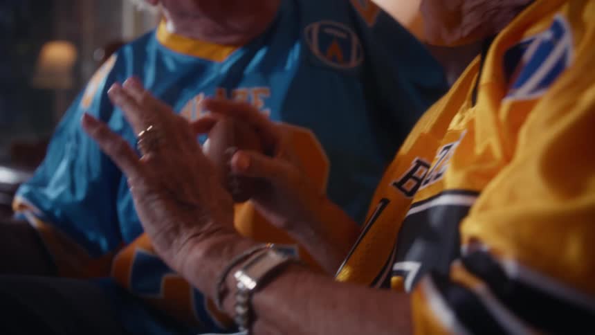 Elderly couple in sports jerseys smiles side by side in lit room with lamp in background