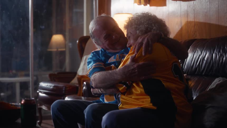 An elderly couple wearing sports jerseys are sitting on a couch and hugging tightly while cheering they are visibly excited and happy likely celebrating a positive moment in a game they are watching