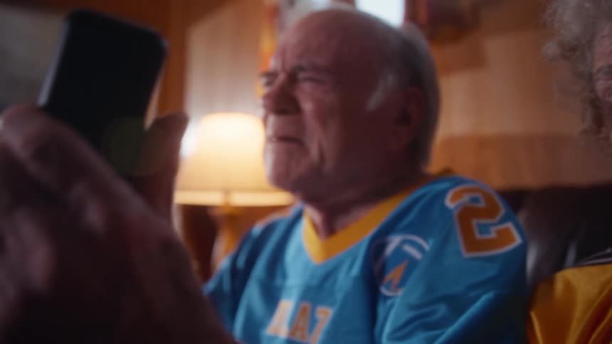 The video opens with a closeup of an elderly woman wearing a yellow football jersey number 1 with blaze written on it she appears distressed or upset wiping her eye the shot then widens to show her seated next to an elderly man wearing a blue jers