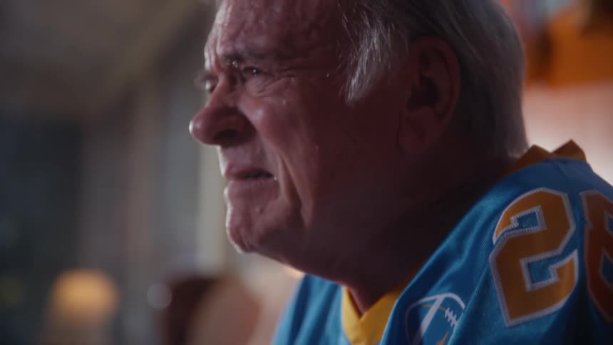 An elderly man sits on his couch in the living room watching a sports game with sadness and disappointment holding the arm of another person beside him