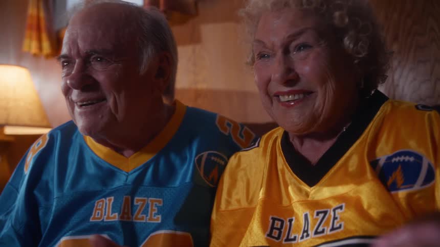 A senior couple wearing matching sports jerseys sits together on a couch watching something with excitement and affection the man is wearing a blue jersey with blaze 99 and the woman is wearing a goldyellow jersey with blaze 77 they are smiling ha