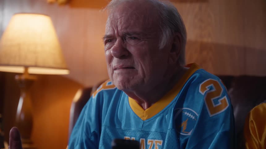 An elderly couple dressed in matching yellow and blue sports jerseys is seated on a brown leather couch in a woodpaneled living room with the man holding a remote controller while watching a game