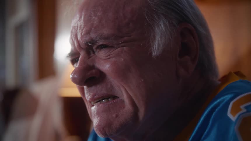 Older man in a blue sports jersey watching something up close in an indoor setting