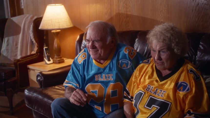 Elderly couple watching television on the couch wearing team jerseys with snacks on the table