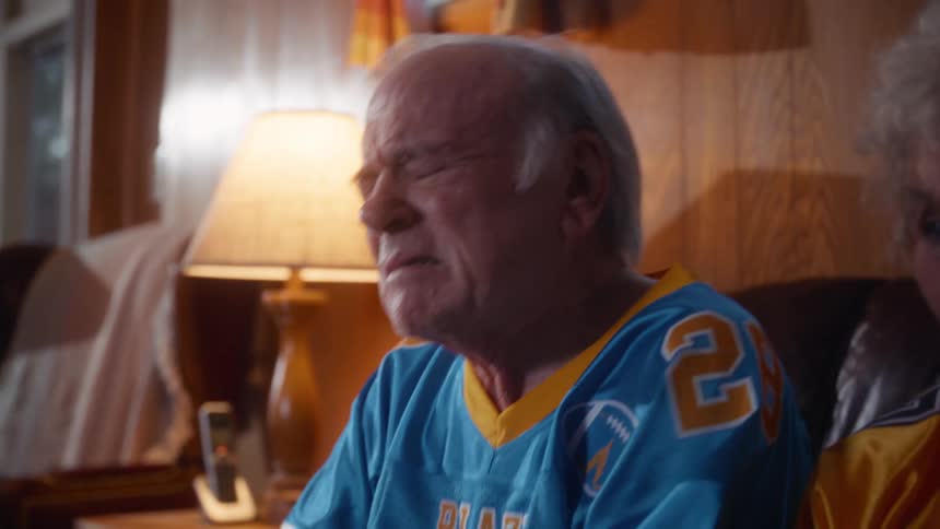 Two elderly caucasian people a man and a woman wearing sports jerseys sitting on a couch and looking forward with excitement then disappointment while watching a game indoors