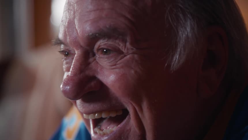 An elderly couple wearing sports jerseys cheers and laughs excitedly while watching a game