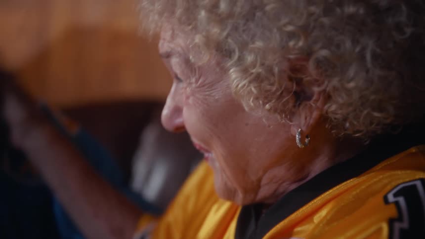 Elderly man and ederly woman wearing football jerseys cheering while laughing and hugging on a couch indoors during night time