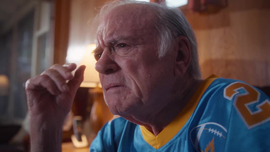 An elderly man sits on a couch in the living room wearing a football jersey watching a sports game with sadness and disappointment