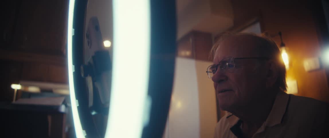 An older man with glasses is visible in a sideprofile view smiling while looking intently at a smartphone camera mounted inside a ring light the reflection of the phone setup and the bright light itself partially obscure the view suggesting he is prep
